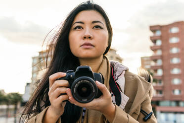 Ethnischer asiatischer weiblicher Fotograf, der ein Foto mit einer professionellen Fotokamera auf einer städtischen Straße schießt - ADSF23959