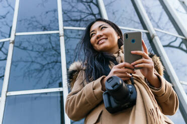Von unten lächelnde asiatische Frau, die mit einer Fotokamera an einer Glaswand stehend in einem Mobiltelefon blättert - ADSF23961