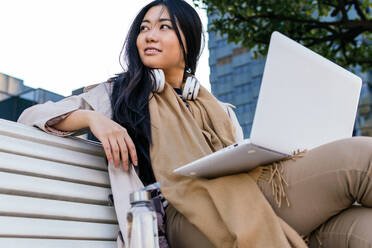 Ethnic young Asian female wearing warm outerwear browsing netbook while working remotely sitting on a bench on the street - ADSF23963