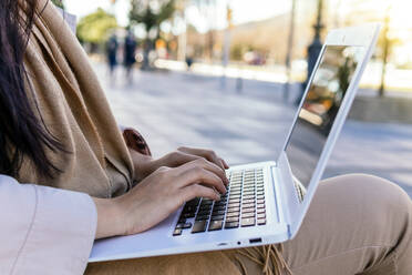 Cropped unrecognizable female wearing warm outerwear browsing netbook while working remotely sitting on a bench on the street - ADSF23964