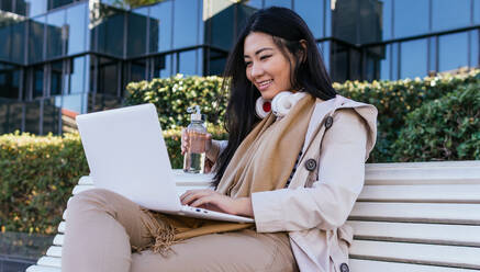 Smiling Asian female in wireless headphones sitting on bench with eco friendly water bottle and browsing netbook while working remotely - ADSF23966