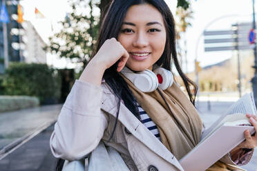 Positive ethnic female in wireless headphones smiling widely while reading book sitting in park and looking at camera - ADSF23967