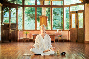 Young mindful male barefoot male sitting in lotus pose practicing yoga on floor near bowl gong and Buddha statuette - ADSF23977