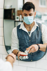 Anonymous male beauty master in sterile mask preparing shave brush with soap in bowl against mirror in bathroom at work - ADSF23992