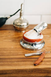 From above of shaving brush with soft bristles in bowl with foamy soap near straight razor on wooden table - ADSF23993