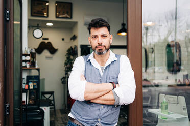 Adult bearded male beauty master with arms crossed looking at camera standing at door of barbershop - ADSF23994