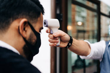 Crop anonymous male hairdresser in sterile mask measuring temperature of colleague with infrared thermometer at door of barbershop - ADSF24001