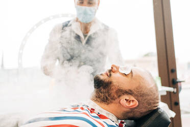 Crop anonymous beauty master in mask near adult bearded male client with cotton pads on closed eyes during vapor procedure in barbershop - ADSF24003