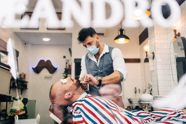 Through glass wall view of anonymous masculine stylist shaving beard of man with cotton pads on eyes using straight razor in beauty salon - ADSF24005