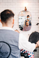 Rückenansicht eines anonymen männlichen Friseurs mit Maske neben einem bärtigen Kunden, der sich im Spiegel eines Friseursalons spiegelt - ADSF24006