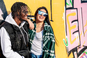 Junger fröhlicher schwarzer Hipster-Mann in der Nähe von zufriedener weiblicher Partnerin mit moderner Sonnenbrille verbringt Zeit zusammen auf der Straße im Sonnenlicht - ADSF24023