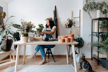 Junge Gärtnerin mit Strohhut, die zu Hause einen Blumenstrauß auf einem Tisch mit verschiedenen Werkzeugen zusammenstellt - ADSF24033