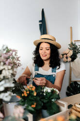 Junge Gärtnerin mit Strohhut, die zu Hause einen Blumenstrauß auf einem Tisch mit verschiedenen Werkzeugen zusammenstellt - ADSF24034
