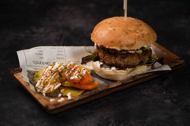 Yummy burger with vegetarian patty and grilled shiitakes between buns near sweet potato and carrot slices with alioli sauce on dark background - ADSF24053