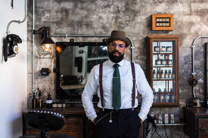 Confident ethnic bearded barber wearing classy outfit standing with hands in pockets in modern barbershop and looking at camera - ADSF24055