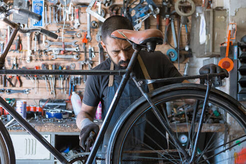 Konzentrierter männlicher Mechaniker mit Bart und Tattoos in Handschuhen repariert Fahrrad in moderner Werkstatt - ADSF24059