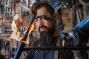 Seriöser bärtiger Mann in Schürze und Schutzhandschuhen mit professioneller Ausrüstung mit Feuer für die Reparatur Fahrrad in der Werkstatt - ADSF24060