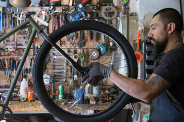 Serious adult man in apron and gloves repairing wheel of bike in modern garage - ADSF24062