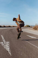 Ganzkörper junger bärtiger Skater in Freizeitkleidung, der springt, während er einen Kickflip auf dem Skateboard auf einer asphaltierten Straße macht - ADSF24064