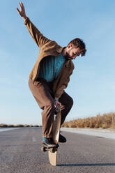 Full body young bearded male skater standing on edge of skateboard keeping balance while performing trick on asphalt road with hand raised and looking down - ADSF24066