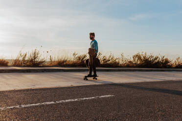 Full body young bearded male riding skateboard with bag and jacket in hand along pavement near asphalt road - ADSF24068