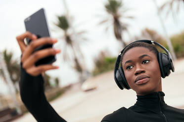 Young ethnic female in casual apparel with wireless headset taking self portrait on mobile phone in a park - ADSF24074