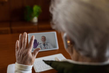 Von oben Rückenansicht einer anonymen älteren Patientin mit TWS-Kopfhörern, die während eines Videoanrufs zu Hause mit einem Arzt auf einem Tablet spricht - ADSF24081