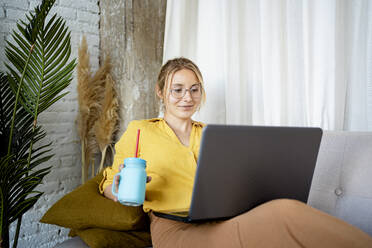Eine Unternehmerin hält ein Einmachglas, während sie zu Hause am Laptop arbeitet - RCPF01038