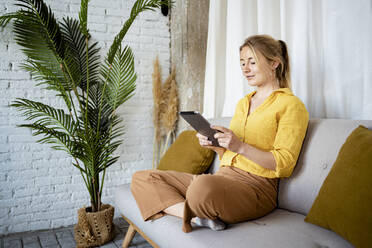 Junge Frau benutzt ein digitales Tablet, während sie zu Hause auf dem Sofa sitzt - RCPF01045