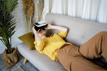 Frau benutzt Virtual-Reality-Simulator, während sie zu Hause auf dem Sofa liegt - RCPF01049
