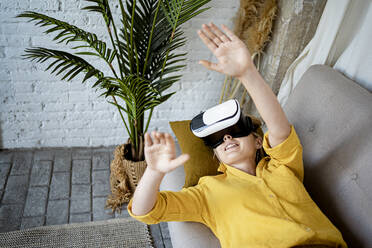 Junge Frau gestikuliert im Virtual-Reality-Simulator, während sie zu Hause auf dem Sofa liegt - RCPF01050