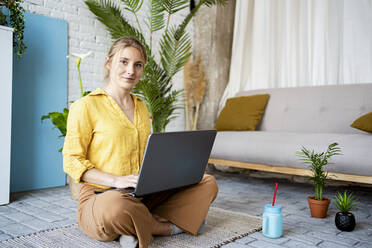 Berufstätige Frau arbeitet am Laptop, während sie zu Hause auf einer Matte sitzt - RCPF01055