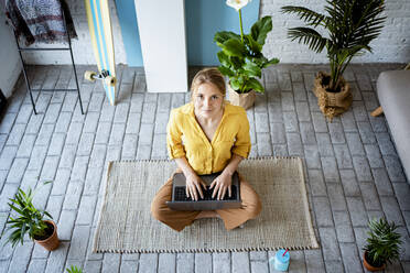 Businesswoman with laptop looking up while sitting on mat at home - RCPF01056