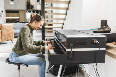 Smiling young woman playing piano in living room at home - MCVF00791