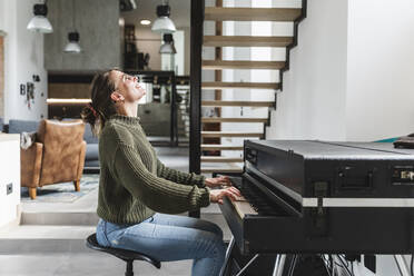 Happy woman playing piano in living room at home - MCVF00793