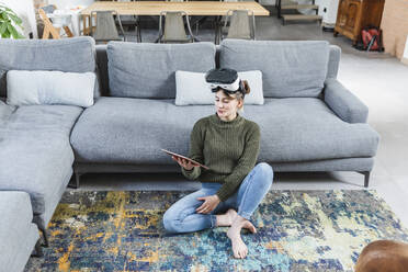 Frau, die ein digitales Tablet hält und ein Virtual-Reality-Headset trägt, sitzt zu Hause - MCVF00797