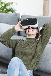 Smiling woman in sweater holding Virtual reality headset at home - MCVF00801