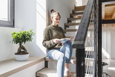 Frau liest ein Buch auf der Treppe zu Hause an einem sonnigen Tag - MCVF00804