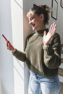 Junge Frau winkt mit der Hand während eines Videogesprächs mit einem Smartphone zu Hause - MCVF00805
