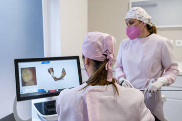 Female dentists wearing surgical cap examining digital image of teeth on screen at clinic - DLTSF01881