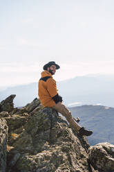 Lächelnder junger Mann mit Hut auf einem Felsen sitzend in den Ferien - RSGF00667