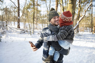 Vater trägt seinen Sohn mit dem Smartphone in der Hand im Winterwald - FVDF00171