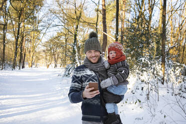 Vater mit Smartphone trägt Sohn im Winter durch den Schnee - FVDF00172