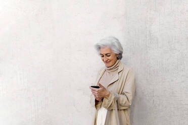 Smiling mature woman using mobile phone in front of wall - ASSF00015