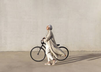 Mature woman walking with bicycle on footpath during sunny day - ASSF00028