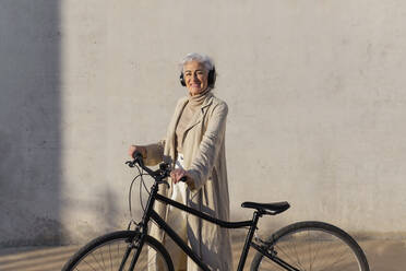 Smiling mature woman wearing headphones standing with bicycle - ASSF00031