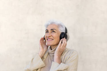 Smiling woman day dreaming while wearing headphones in front of wall - ASSF00033