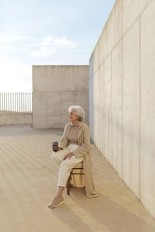 Frau mit wiederverwendbarem Kaffeebecher, die auf einem Hocker sitzt und wegschaut - ASSF00045