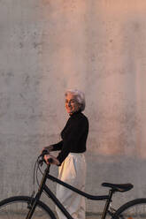 Smiling woman standing with bicycle by wall - ASSF00057