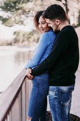 Lächelnder Mann umarmt seine Freundin vor einem Geländer - EBBF03437
